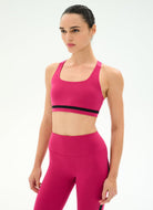 A woman modeling a bright pink activewear set consisting of a scoop-neck sports bra with a black band detail and matching high-waisted leggings, shot against a plain white studio background.