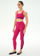 A woman modeling a bright pink activewear set consisting of a sports bra with a black band and matching high-waisted leggings with black side stripes, paired with black flats, against a plain white studio background.