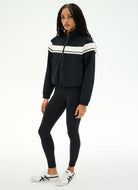 A woman modeling a black athletic zip-up jacket with white chest stripes, paired with black leggings and white sneakers, against a plain white studio background.