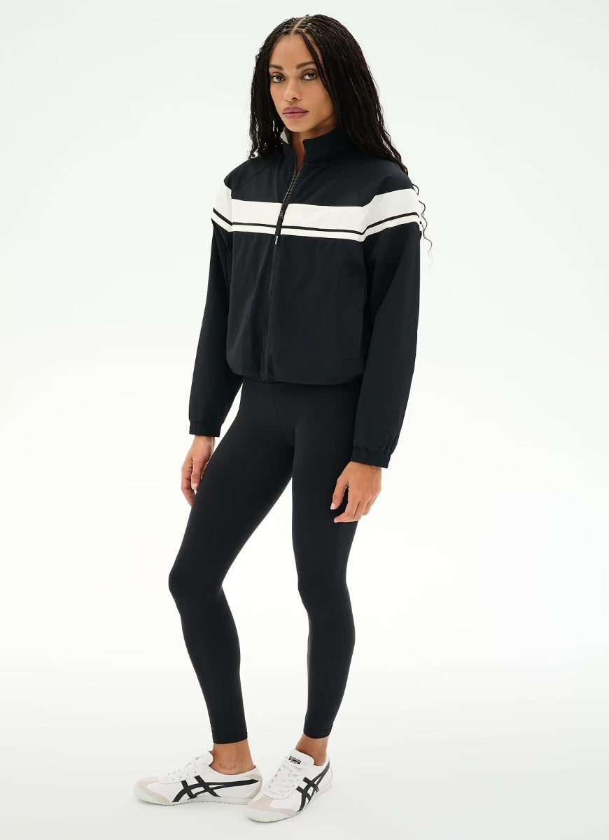 A woman modeling a black athletic zip-up jacket with white chest stripes, paired with black leggings and white sneakers, against a plain white studio background.
