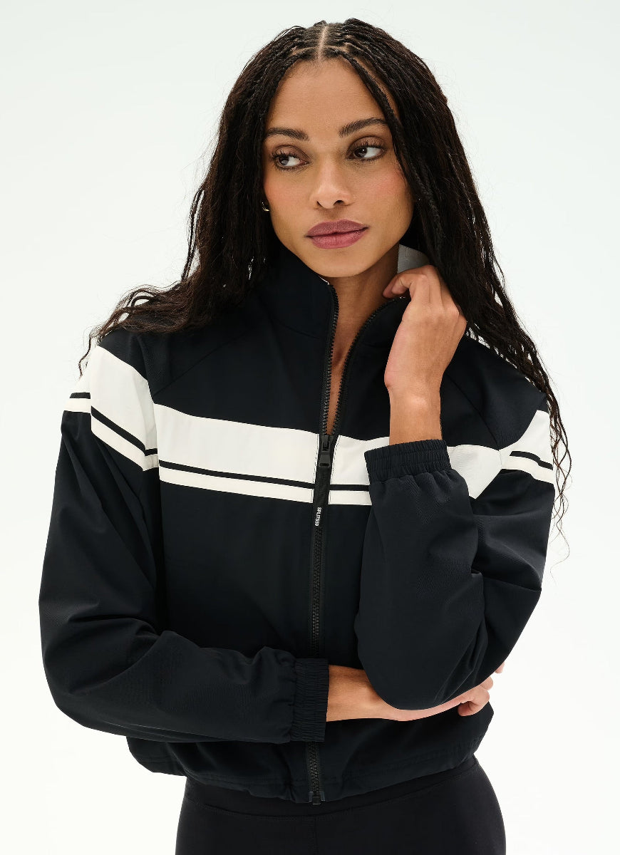 A woman modeling a black zip-up track jacket with bold white stripe accents across the chest and shoulders, set against a plain white studio background.