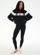 A woman modeling a black zip-up track jacket with white shoulder stripes, paired with black leggings and white sneakers, against a plain white studio background.