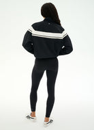 A woman viewed from behind wearing a black cropped track jacket with white stripe accents on the shoulders, black leggings, and white sneakers, against a plain white studio background.