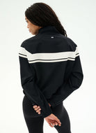A woman shown from the back wearing a black athletic jacket with two horizontal white stripes across the upper back and shoulders, paired with black leggings, against a plain white studio background.