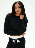 A woman modeling a black long-sleeve cropped sweatshirt with a white ribbed waistband, paired with black sweatpants featuring a white drawstring, against a plain white studio background.
