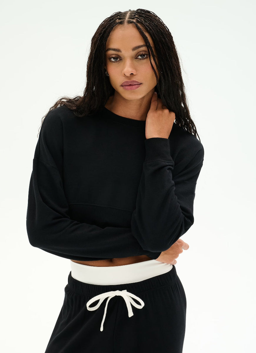 A woman modeling a black long-sleeve cropped sweatshirt with a white ribbed waistband, paired with black sweatpants featuring a white drawstring, against a plain white studio background.