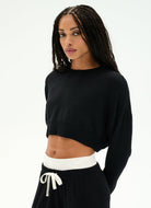 A woman modeling a black long-sleeve cropped sweatshirt with a wide white waistband, paired with black drawstring sweatpants, against a plain white studio background.