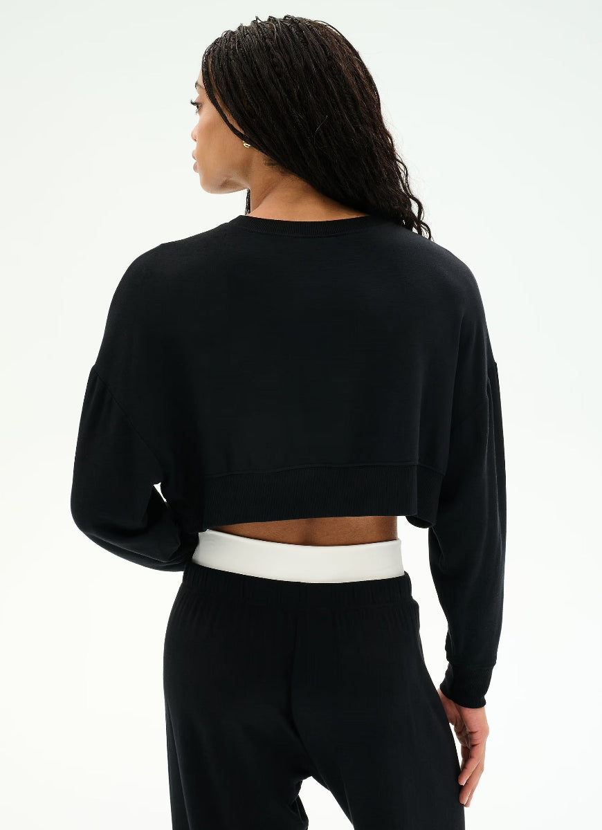 A woman shown from the back wearing a black long-sleeve cropped sweatshirt and black pants with a white waistband, against a plain white studio background.