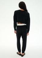 A woman shown from the back wearing a black long-sleeve cropped top with a white underlayer peeking out at the waist, paired with black jogger pants and black flats, against a plain white studio background.
