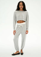 A woman modeling a light gray two-piece loungewear set consisting of a cropped long-sleeve sweatshirt and matching jogger pants with a white drawstring waistband, paired with black flats against a white studio background.