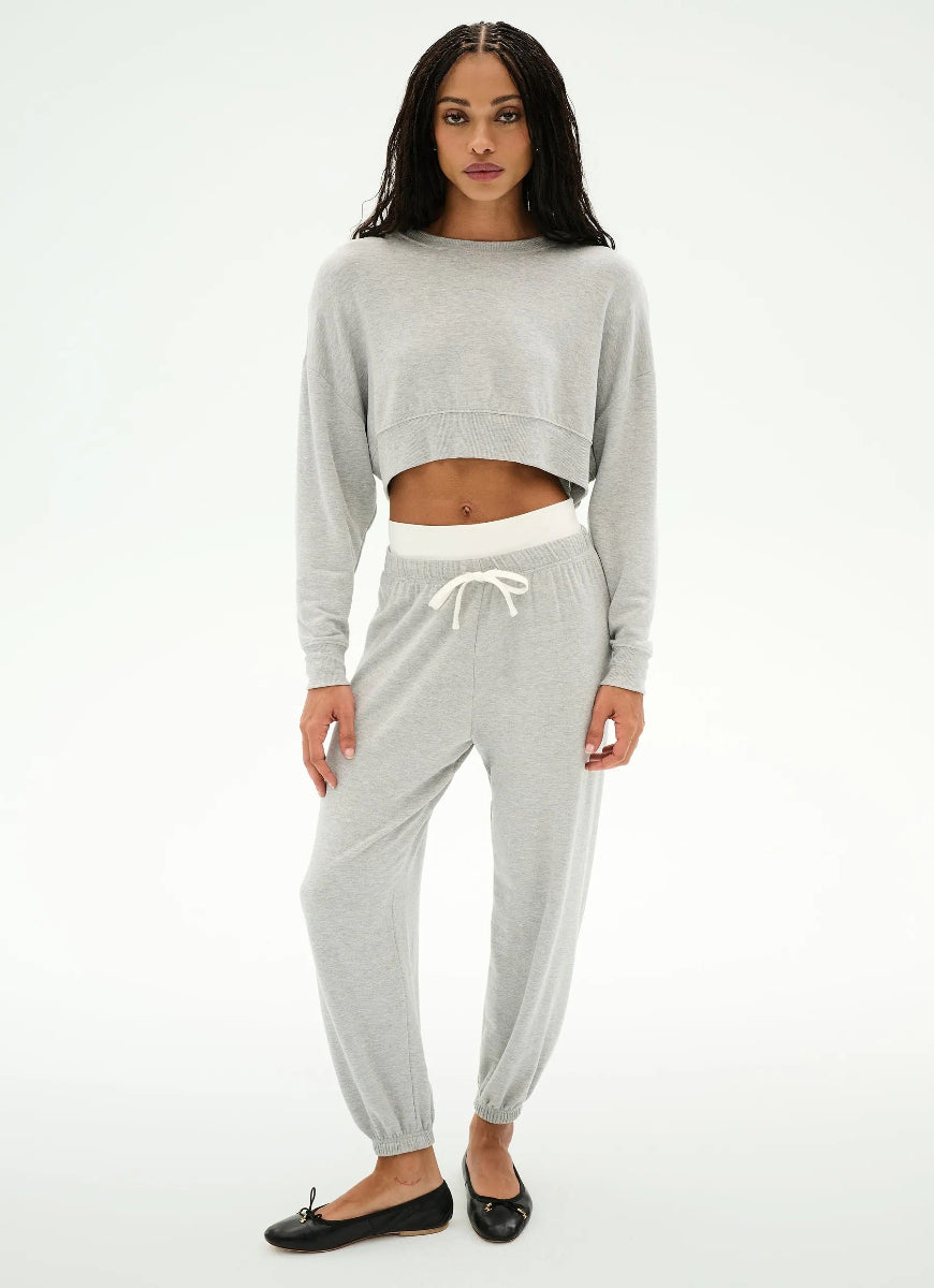 A woman modeling a light gray two-piece loungewear set consisting of a cropped long-sleeve sweatshirt and matching jogger pants with a white drawstring waistband, paired with black flats against a white studio background.