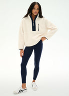 A woman modeling a cream-colored quarter-zip fleece pullover with navy blue trim, paired with navy blue leggings and white sneakers, against a plain white studio background.