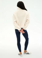 A woman shown from the back wearing a cream-colored fuzzy mock-neck sweater, dark navy leggings, and white sneakers with black stripes, set against a plain white studio background.