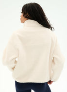 A woman shown from the back wearing a cream-colored sherpa fleece pullover jacket and dark navy leggings against a plain white studio background.