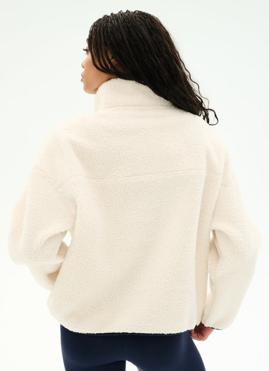 A woman shown from the back wearing a cream-colored sherpa fleece pullover jacket and dark navy leggings against a plain white studio background.