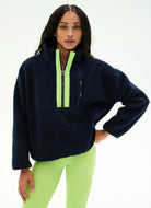 A woman modeling a dark navy blue fleece pullover with bright neon green contrast trim along the half-zip collar and a matching neon green zippered pocket on the chest, paired with neon green leggings against a plain white studio background.