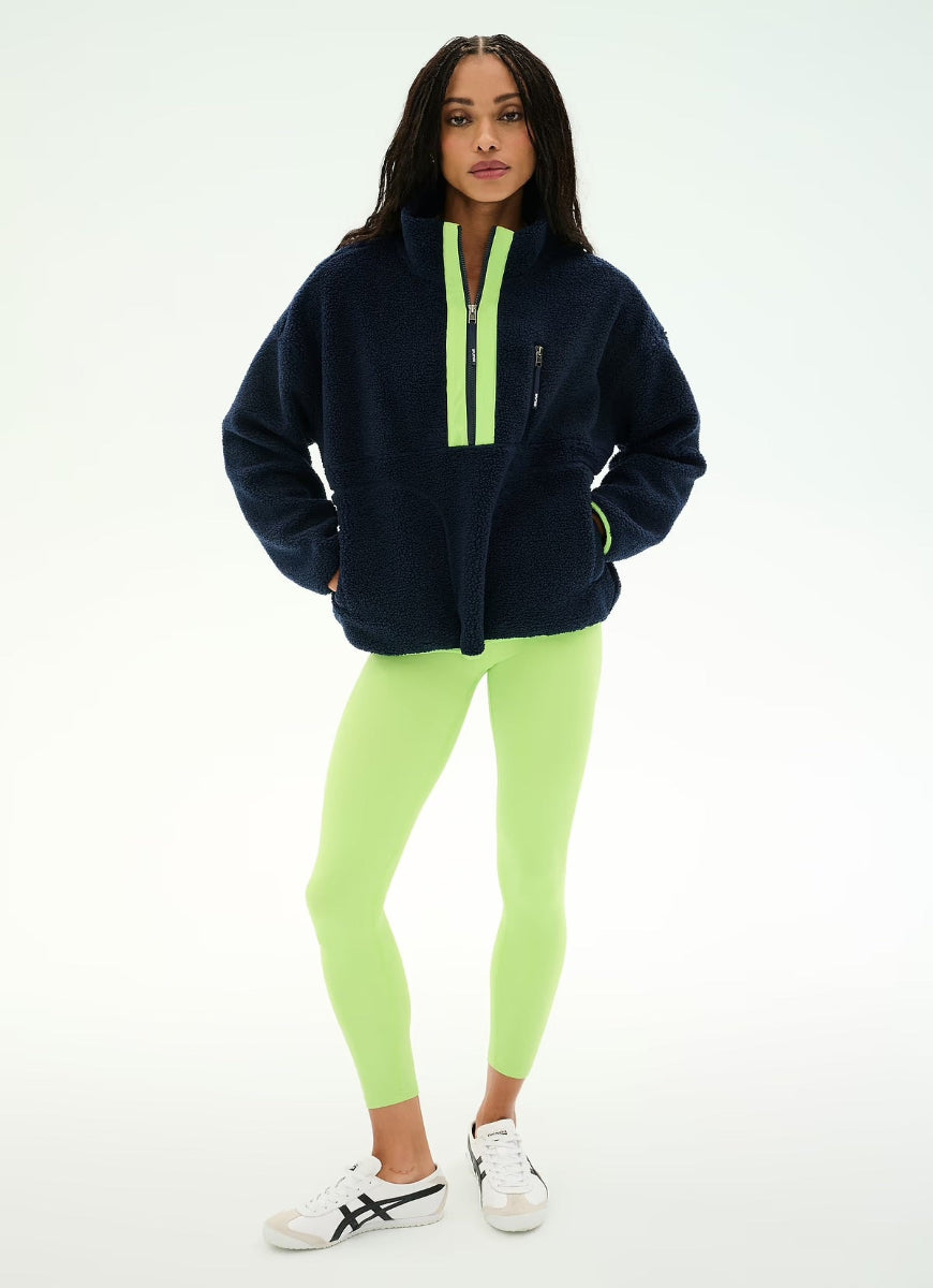 A woman modeling a navy blue fleece pullover with a bright neon green zipper placket, paired with neon green leggings and white sneakers, against a plain white studio background.