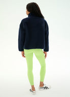 A woman shown from the back wearing a dark navy fleece pullover, neon lime green leggings, and white sneakers with black stripes, set against a plain white studio background.