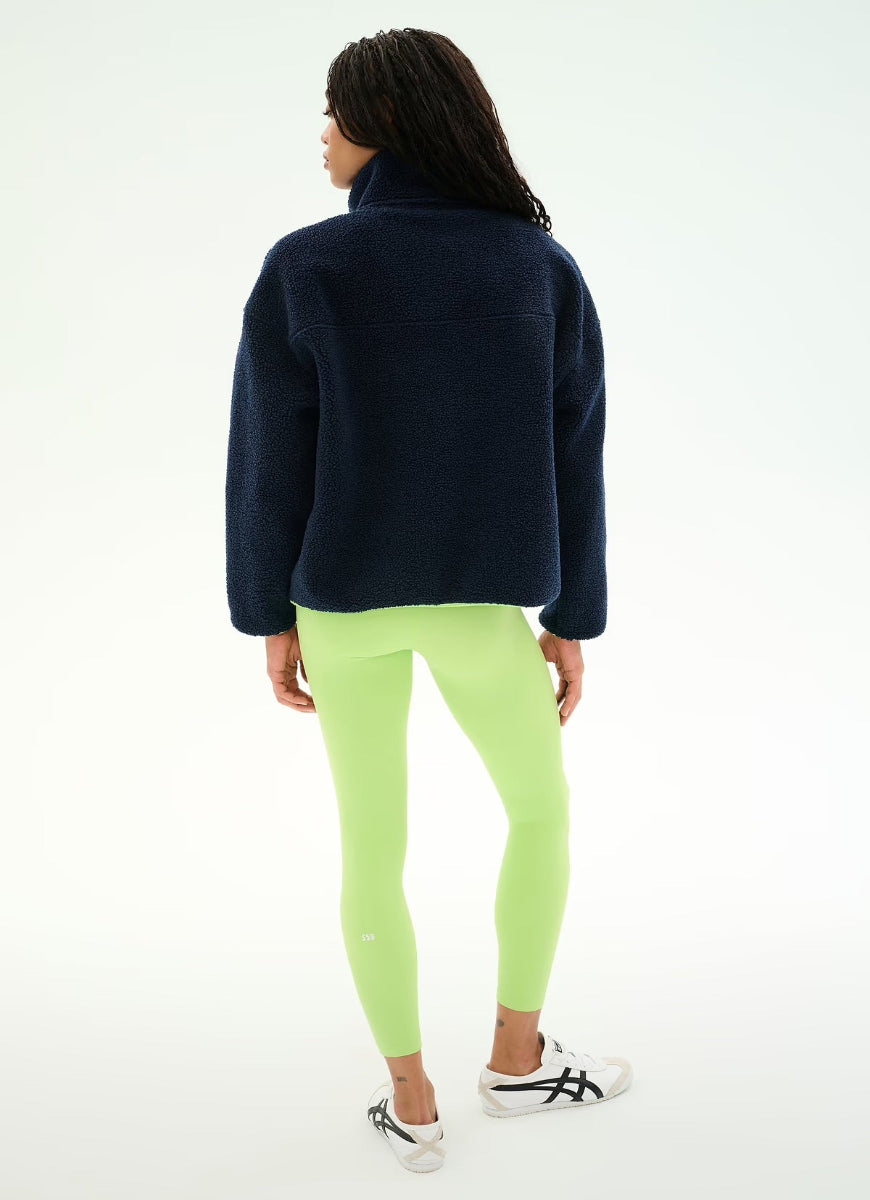 A woman shown from the back wearing a dark navy fleece pullover, neon lime green leggings, and white sneakers with black stripes, set against a plain white studio background.