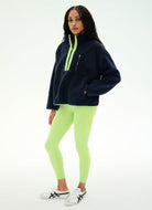A woman modeling a navy blue fleece pullover with a bright neon green half-zip collar and zipper details, paired with neon green leggings and white sneakers with black stripes, against a plain white studio background.