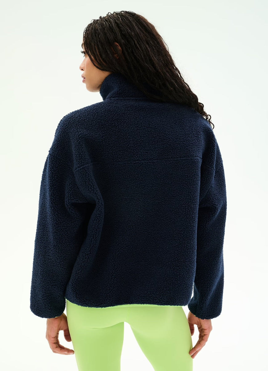 A woman shown from the back wearing a dark navy blue sherpa fleece pullover and neon green leggings against a clean white studio background.