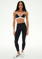 A woman modeling a black and white athletic set consisting of a strappy triangle sports bra and high-waisted black leggings, paired with white sneakers, against a plain white studio background.