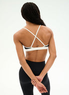 A woman shown from the back wearing a white sports bra with crisscross straps and black leggings against a plain white studio background.