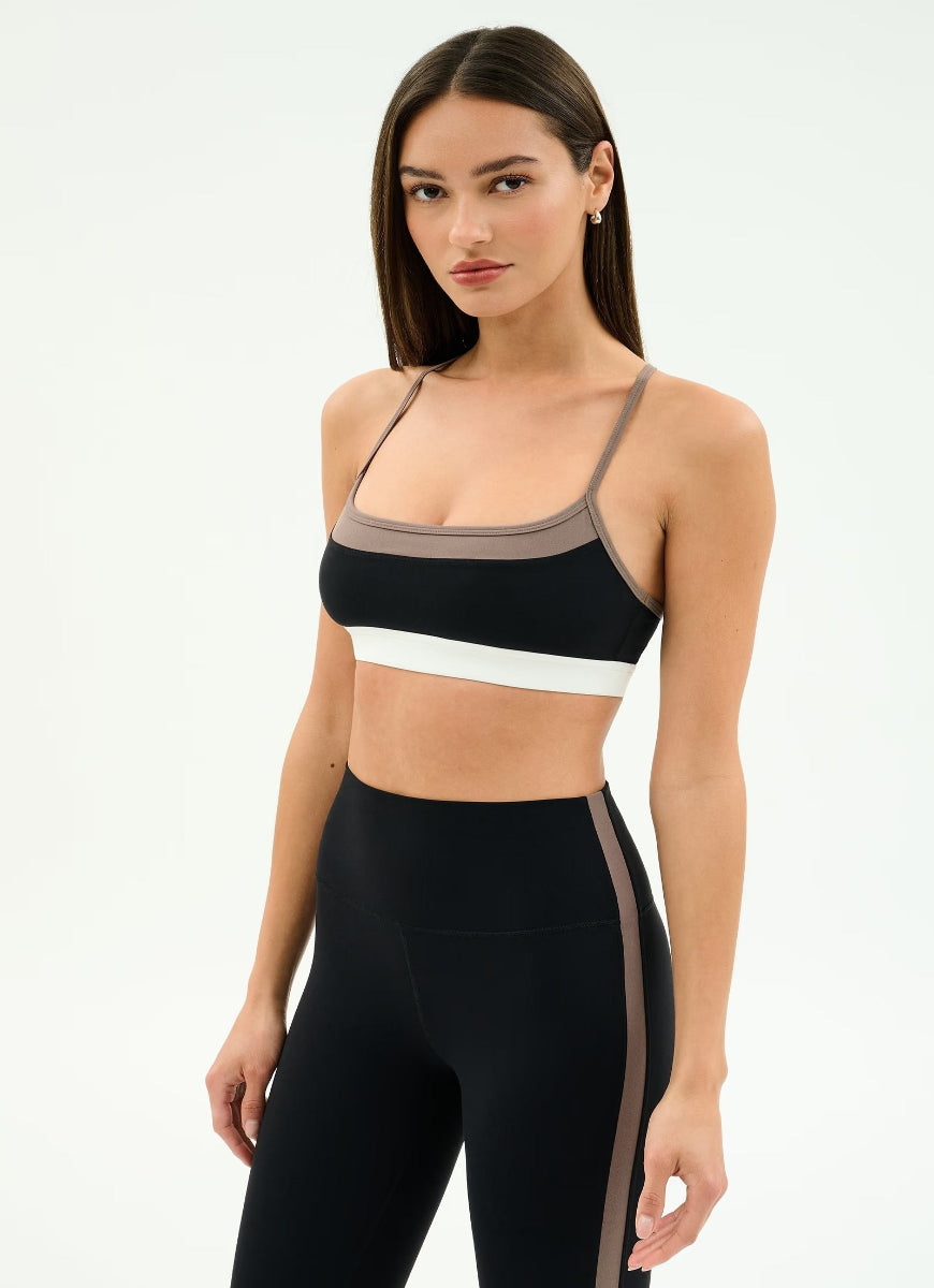 Woman wearing a black sports bra with beige and white accents and black leggings on a light gray background